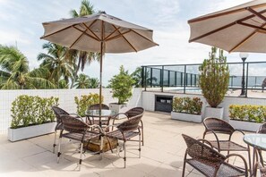 An outdoor patio with wicker furniture, umbrellas, and potted plants. - Bright Suite Unforgettable Beach Retreats - Beachfront with Rooftop Pool (Paraíba)