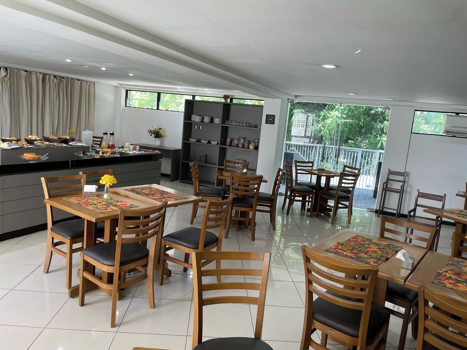 A modern dining area with wooden tables and chairs, a kitchen in the background, and large windows offering a view of greenery.