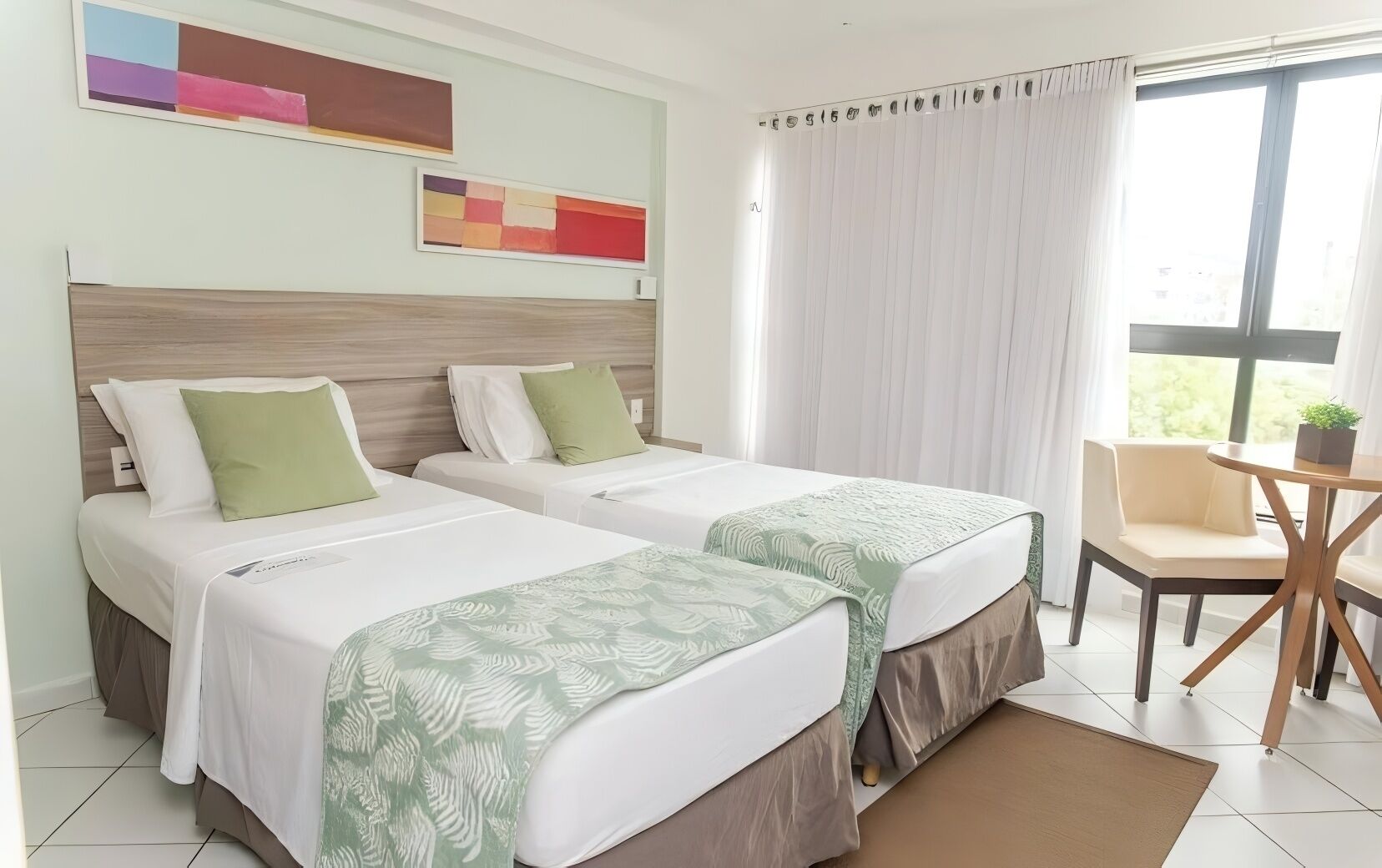 Quarto de hotel com duas camas, uma mesinha e uma cadeira.