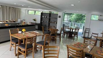 Uma sala de jantar moderna com mesas e cadeiras de madeira, uma cozinha ao fundo e amplas janelas com vista para o verde.