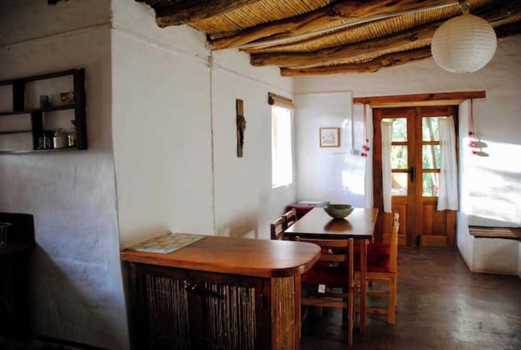 A rustic dining area with a wooden table and chairs, a shelf with jars, and a door leading outside.