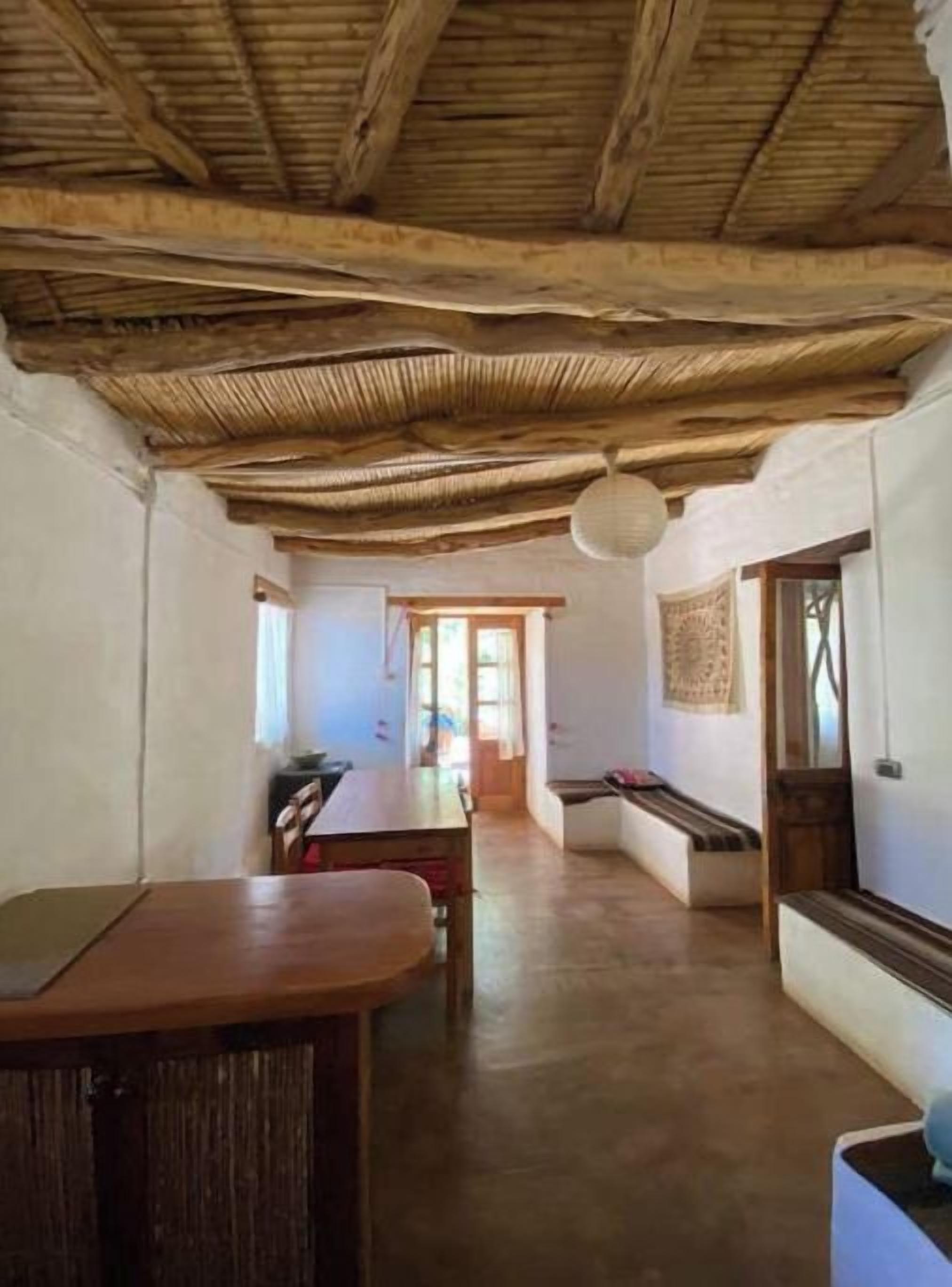 A rustic interior with a wooden ceiling, a dining area with a bench, and a view through a doorway.