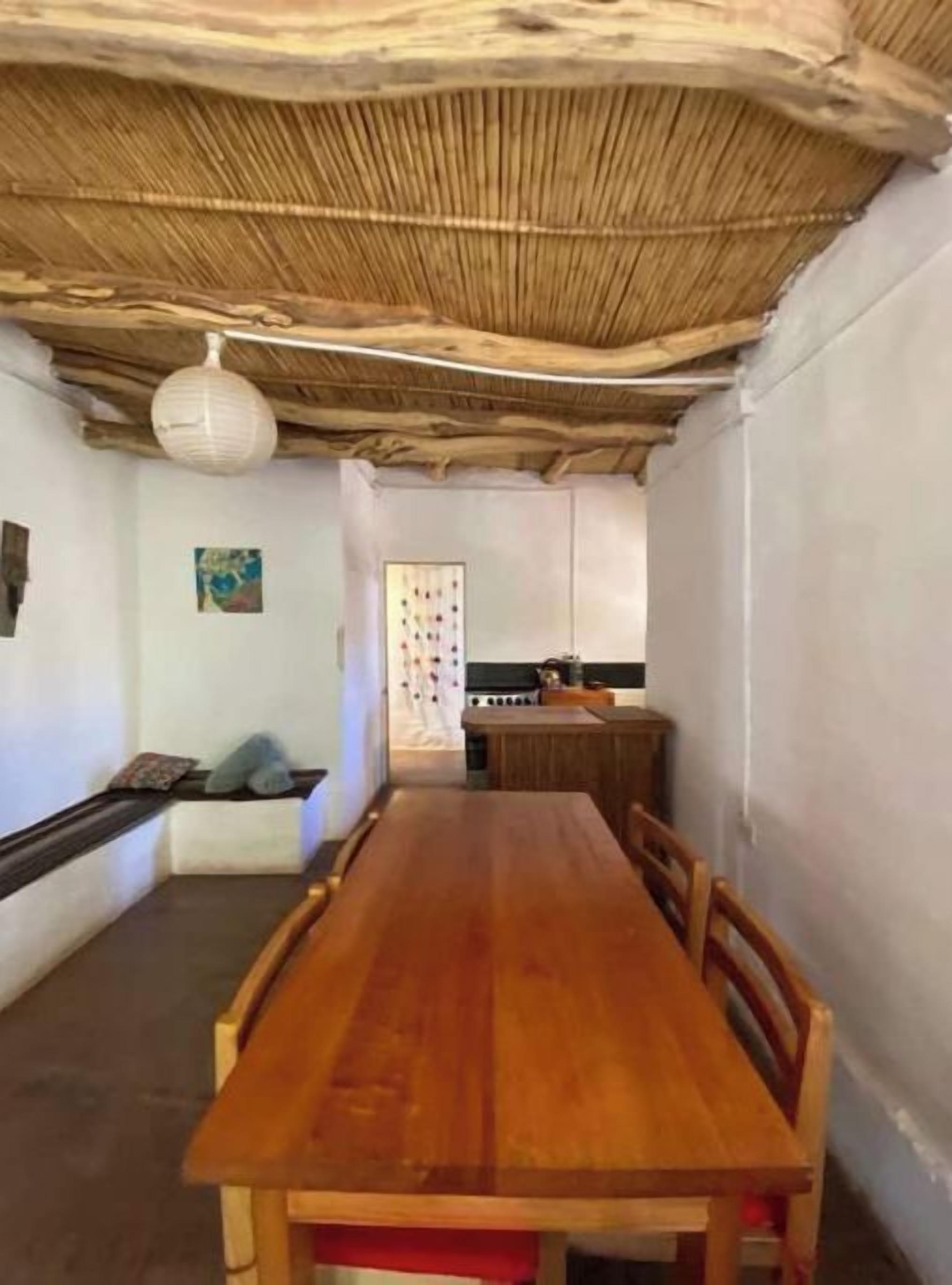 A room with a wooden dining table, chairs, a thatched ceiling, and a hanging light fixture.