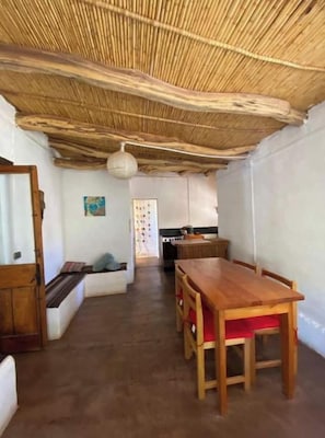 A room with a thatched ceiling, a wooden dining table with red cushions, and a kitchen area in the background.