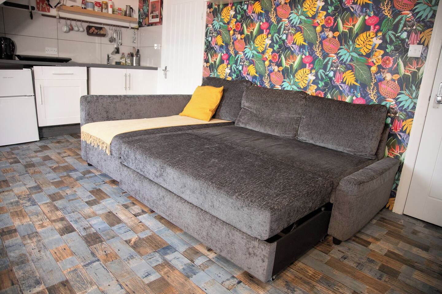 A grey sofa with a yellow throw pillow in a room with a patterned wallpaper and a checkered wooden floor.