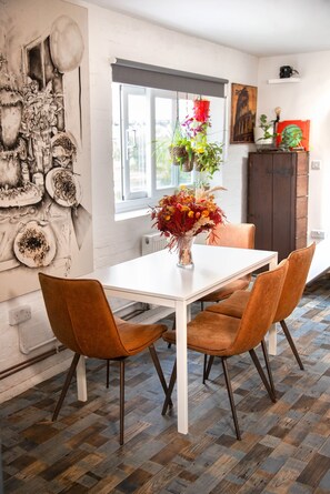 A dining area with a white table and two brown chairs, a vase with flowers, and a wall with various decorations. - 3 Bed Flat - 1 Min Walk Underground (London)