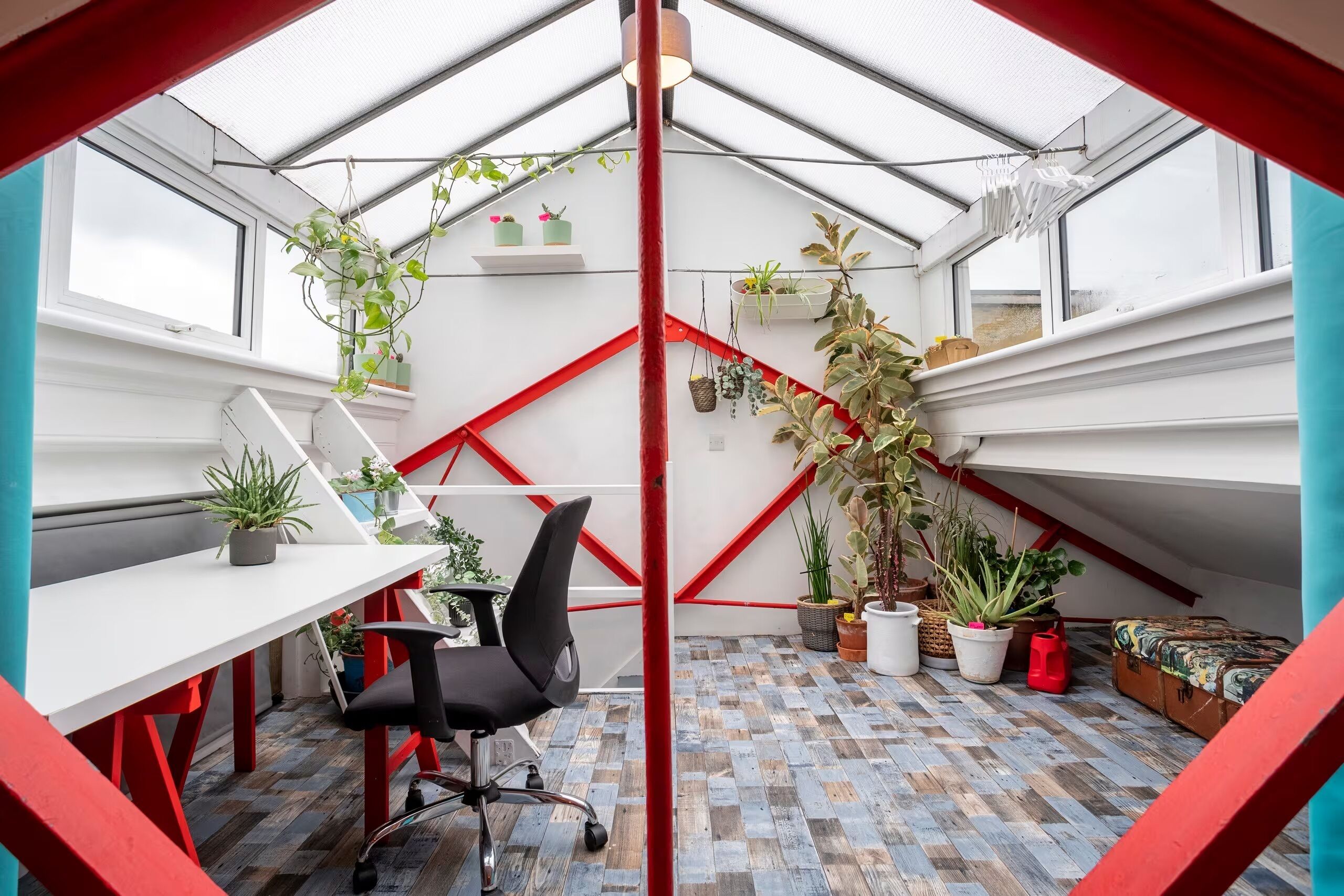 Una habitación moderna y compacta con techo inclinado, un escritorio blanco, una silla de oficina negra y varias plantas en macetas.