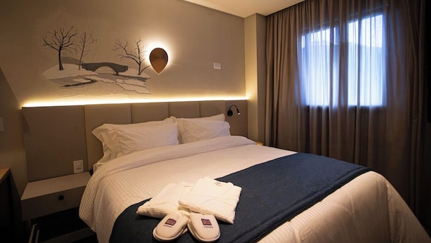 Habitación de hotel con cama, mesita de noche, una obra de arte en la pared y ventana con cortinas.
