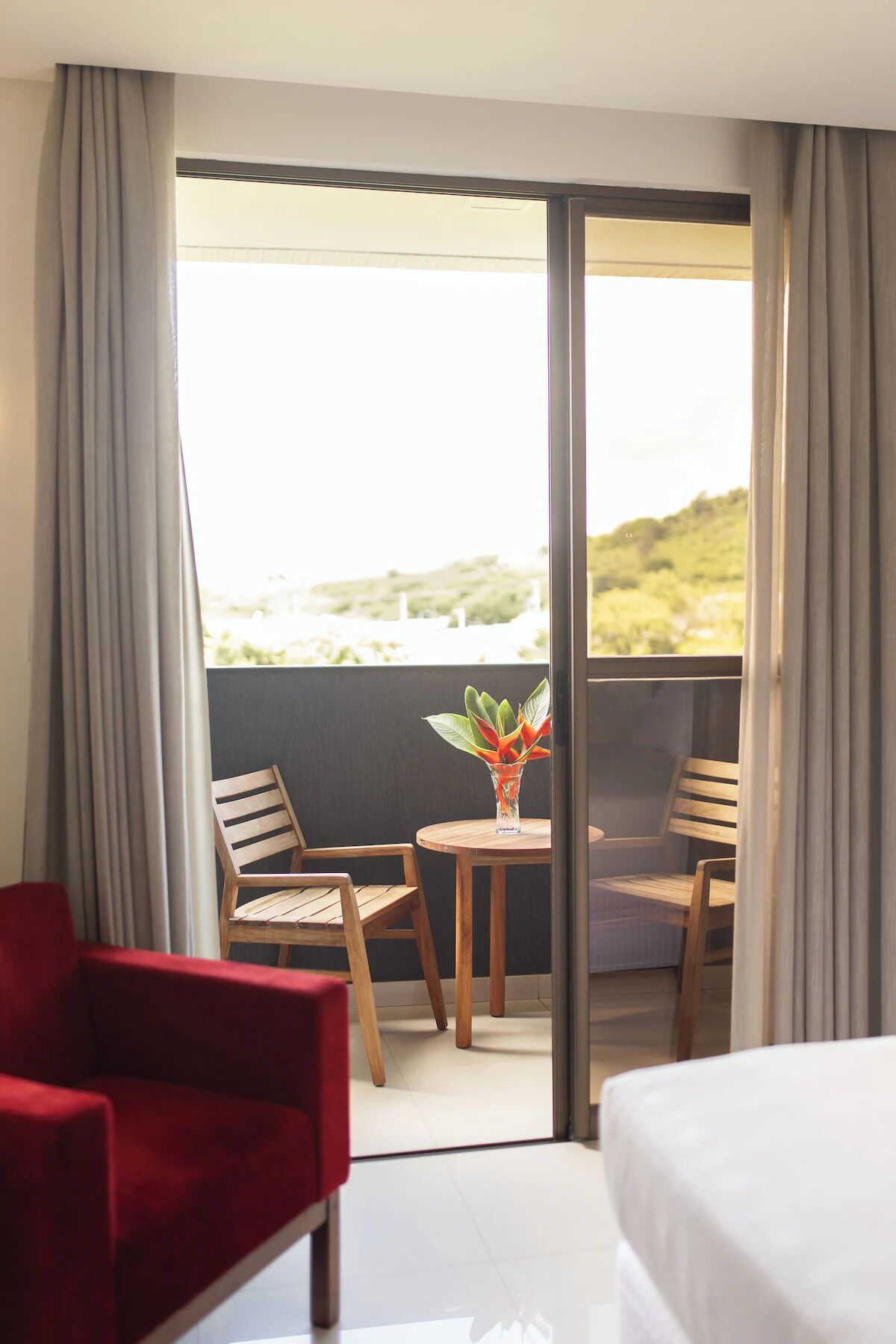 Quarto de hotel com varanda, uma cadeira vermelha, mesa de madeira com um vaso de flores e uma cama.