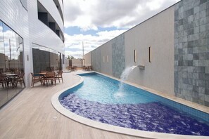 En inomhuspool med en böjd design, omgiven av träbord och stolar, och en modern byggnad med stora fönster och en vägg beklädd med kakel.