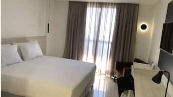 Quarto de hotel com uma cama grande, uma escrivaninha com cadeira, uma luminária de mesa e uma janela com cortinas.