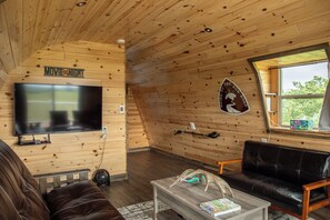 L’intérieur d’un chalet accueillant, avec un téléviseur fixé au mur, un canapé en cuir, une table basse en bois et une fenêtre donnant sur la forêt.