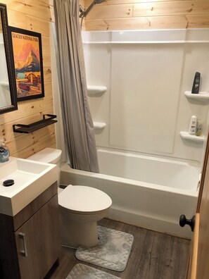A bathroom with a toilet, sink, and bathtub. There is a shower curtain, a framed picture on the wall, and a small shelf with toiletries. - Glacier Aurora - Stargazing Wonder overlooking the Forest and the East Rockies (Babb)