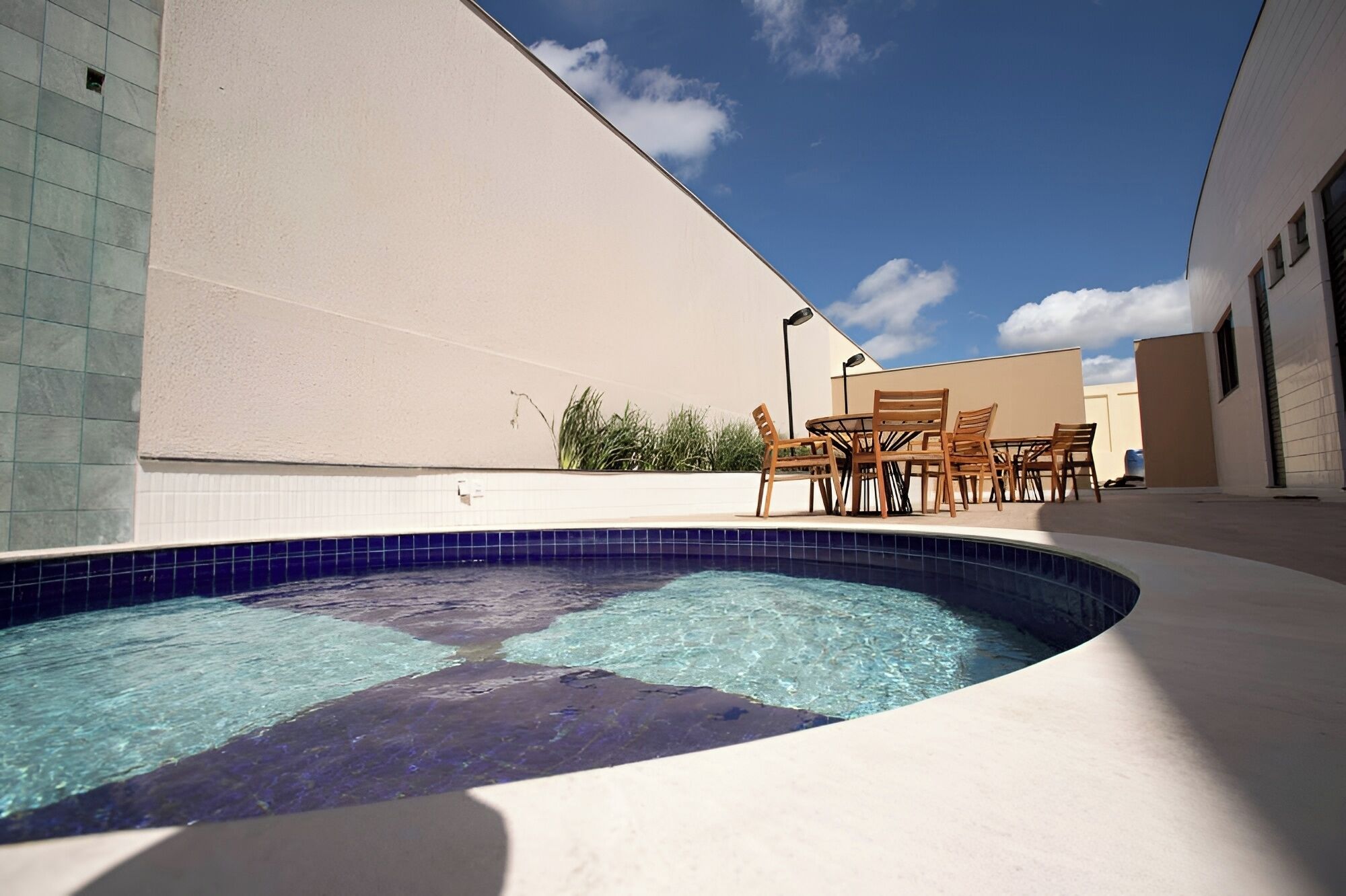 Piscina com borda curva, rodeada por um muro bege e área revestida com azulejos. Uma mesa e cadeiras de madeira estão dispostas perto.