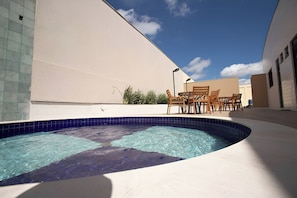 A swimming pool with a curved edge, surrounded by a beige wall and a tiled area. There is a wooden table and chairs set up nearby.