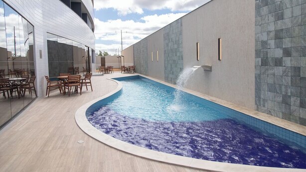 An indoor swimming pool with a curved design, surrounded by wooden tables and chairs, and a modern building with large windows and a tiled wall. - Unwind in a Bright Space with Balcony & Buffet Breakfast Near Local Attractions (Pernambuco)
