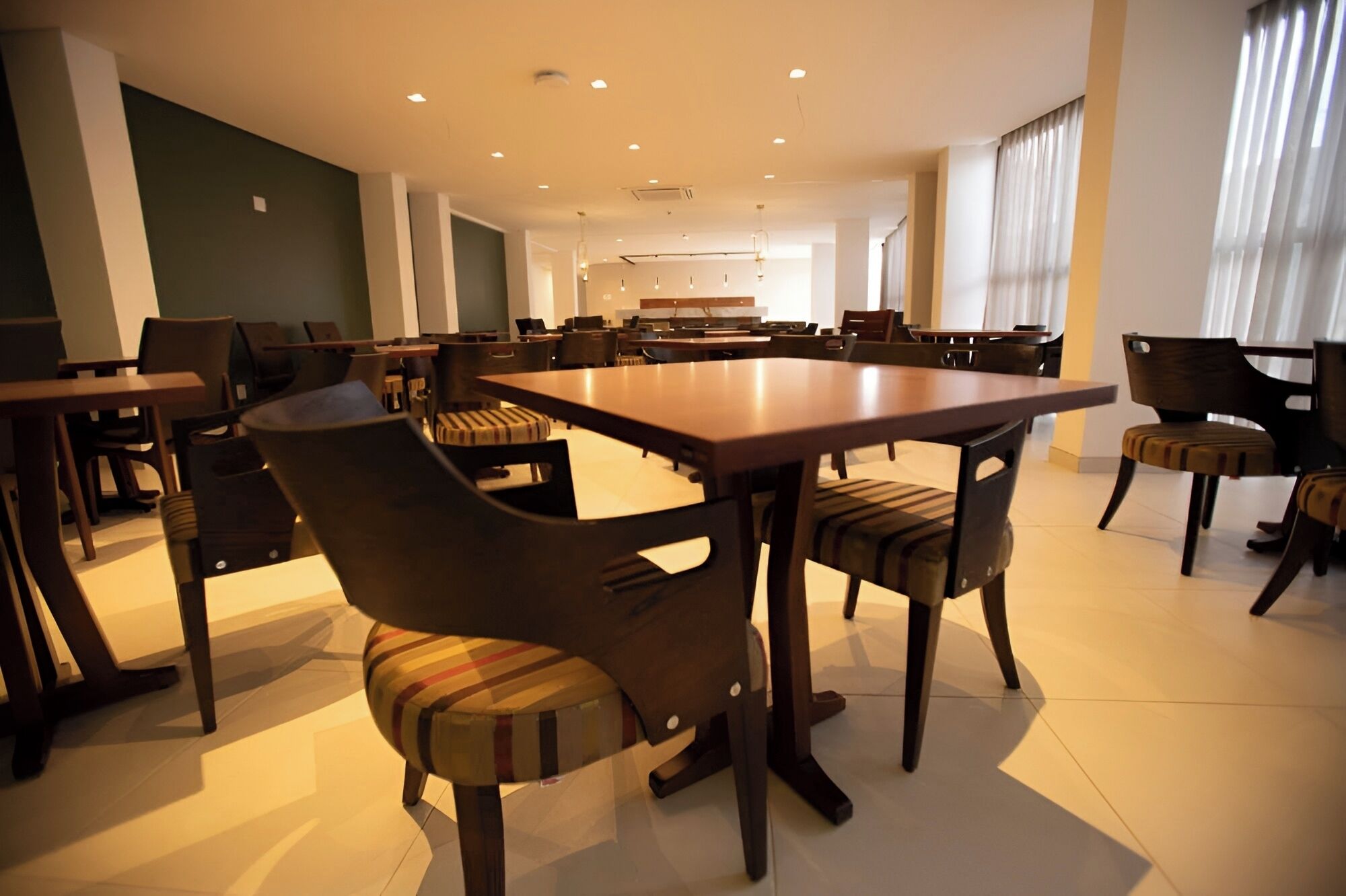 Uma sala de jantar espaçosa com várias mesas e cadeiras, uma cozinha ao fundo e grandes janelas que permitem entrada de luz natural.