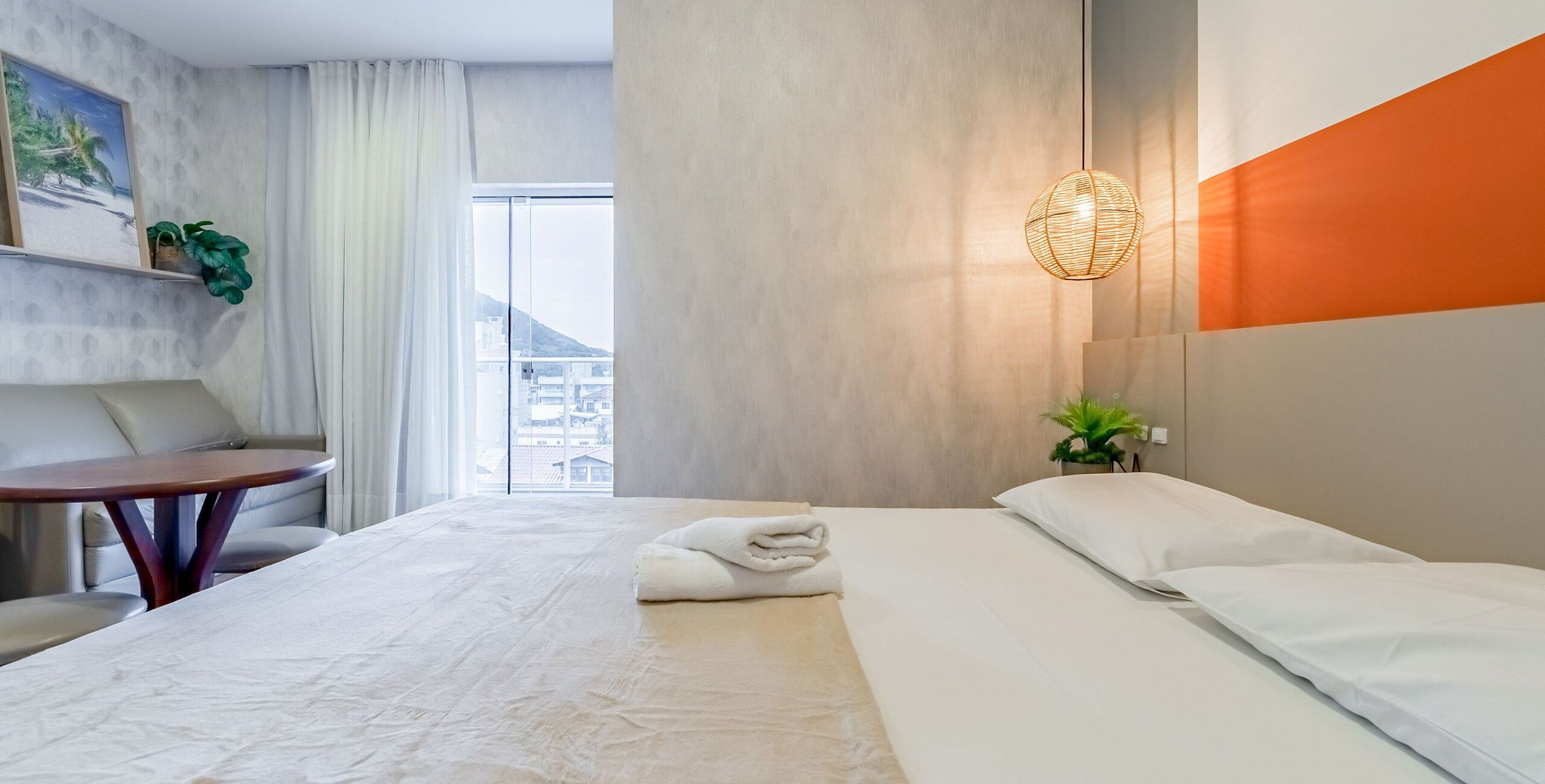 Habitación de hotel con cama, escritorio, silla, ventana con vista a edificios y lámpara de pared.