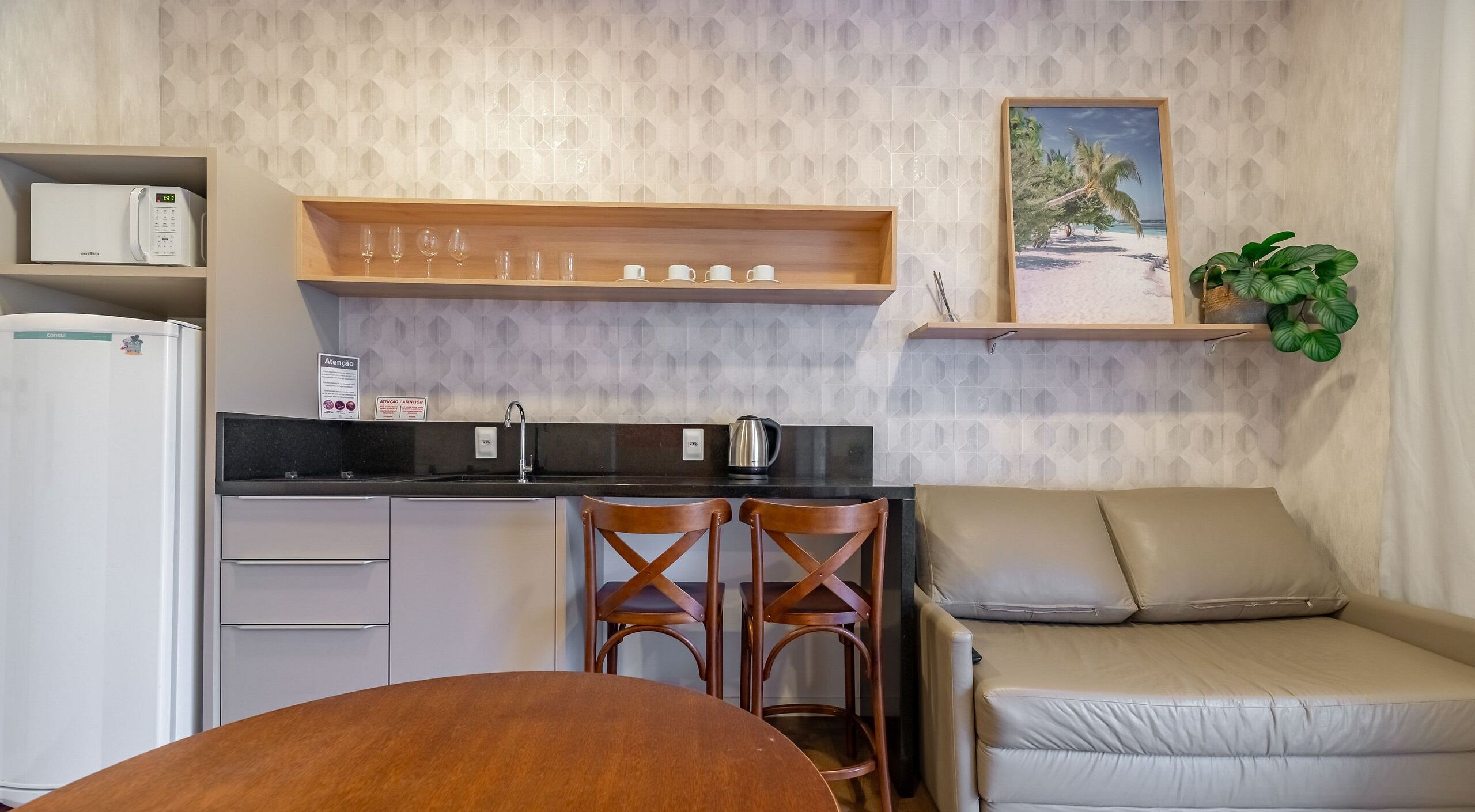 A compact kitchen with a refrigerator, a coffee maker, and a kettle. There are two wooden chairs and a sofa with cushions. A shelf above the counter holds cups and a plant.