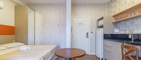 Habitación de hotel pequeña y funcional con cama, una mesita redonda, kitchenette y una puerta que da a otra habitación.