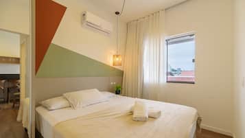 A hotel room with a bed, a window with curtains, an air conditioning unit, and a plant on the nightstand.