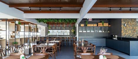 A restaurant with wooden tables and chairs, a bar area with shelves of bottles, and a green hanging plant.