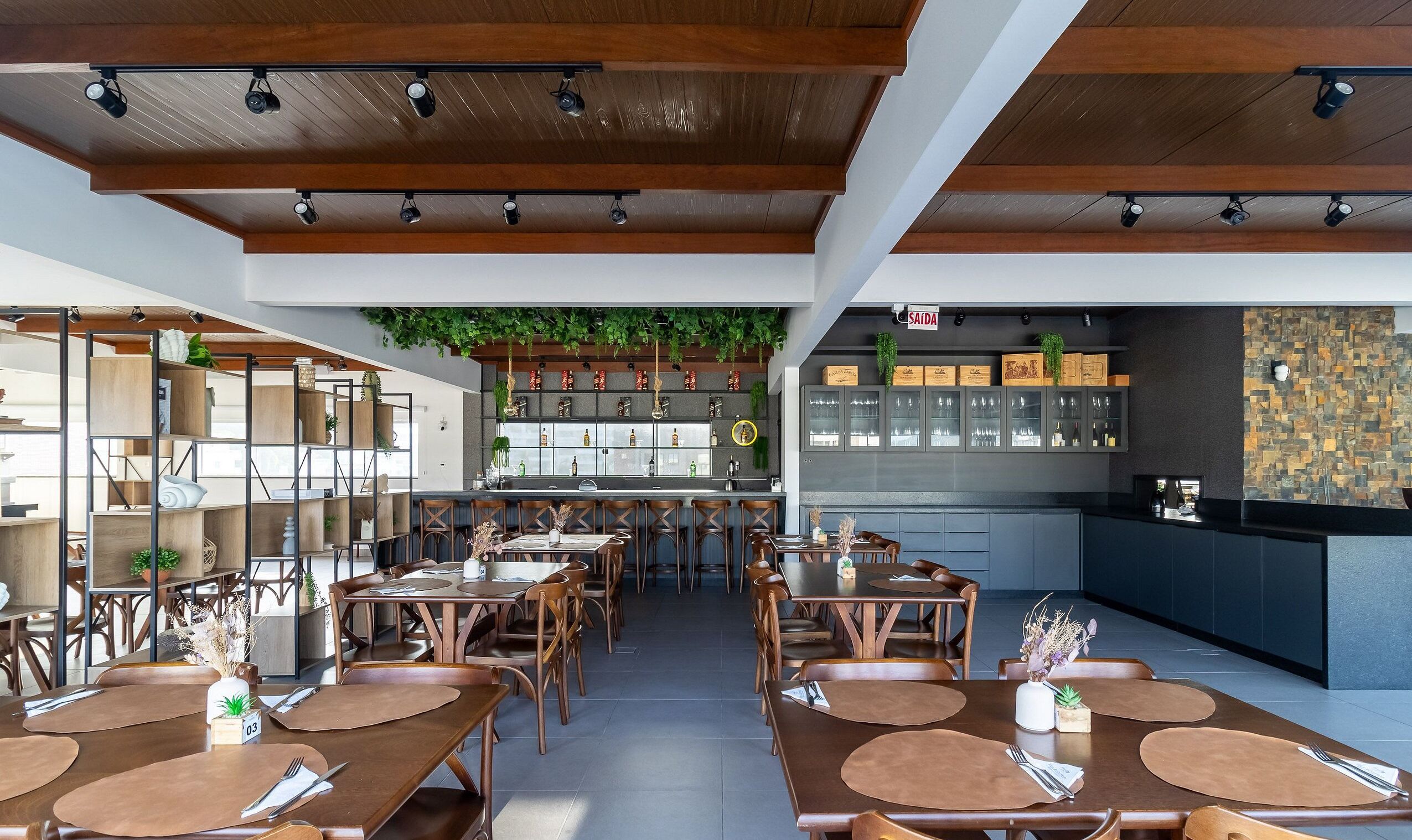 A restaurant interior with wooden tables and chairs, a bar area with shelves of bottles, and a green hanging plant.