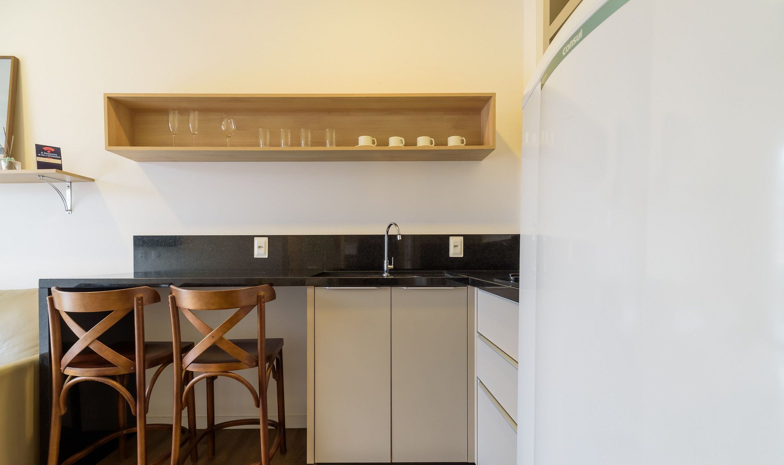 Une cuisine avec un plan de travail noir, deux tabourets de bar en bois, une étagère en bois au-dessus du plan de travail où sont rangés des verres et des tasses, et un réfrigérateur blanc.