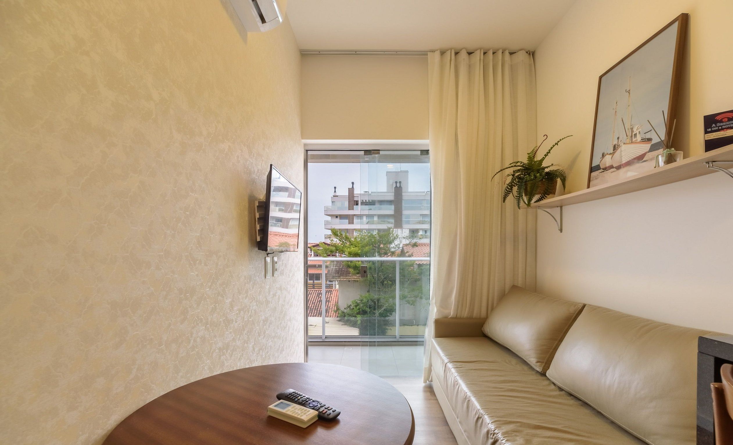 A hotel room with a sofa, a small table with a remote control, a potted plant, and a window with a view of buildings.