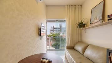 A hotel room with a sofa, a small table with a remote control, a potted plant, and a window with a view of buildings.