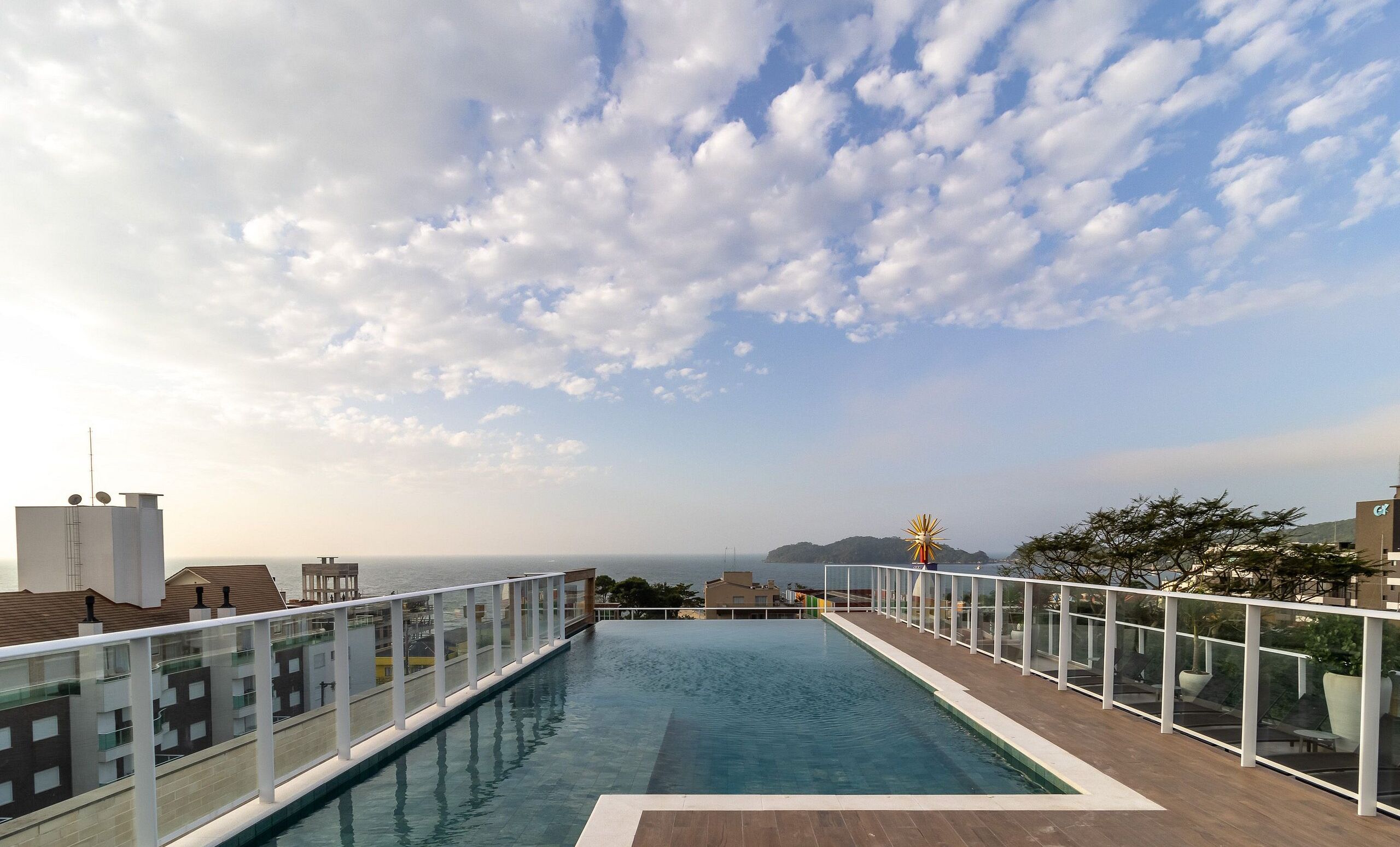 A rooftop infinity pool with a clear view of the sea and distant land under a partly cloudy sky.