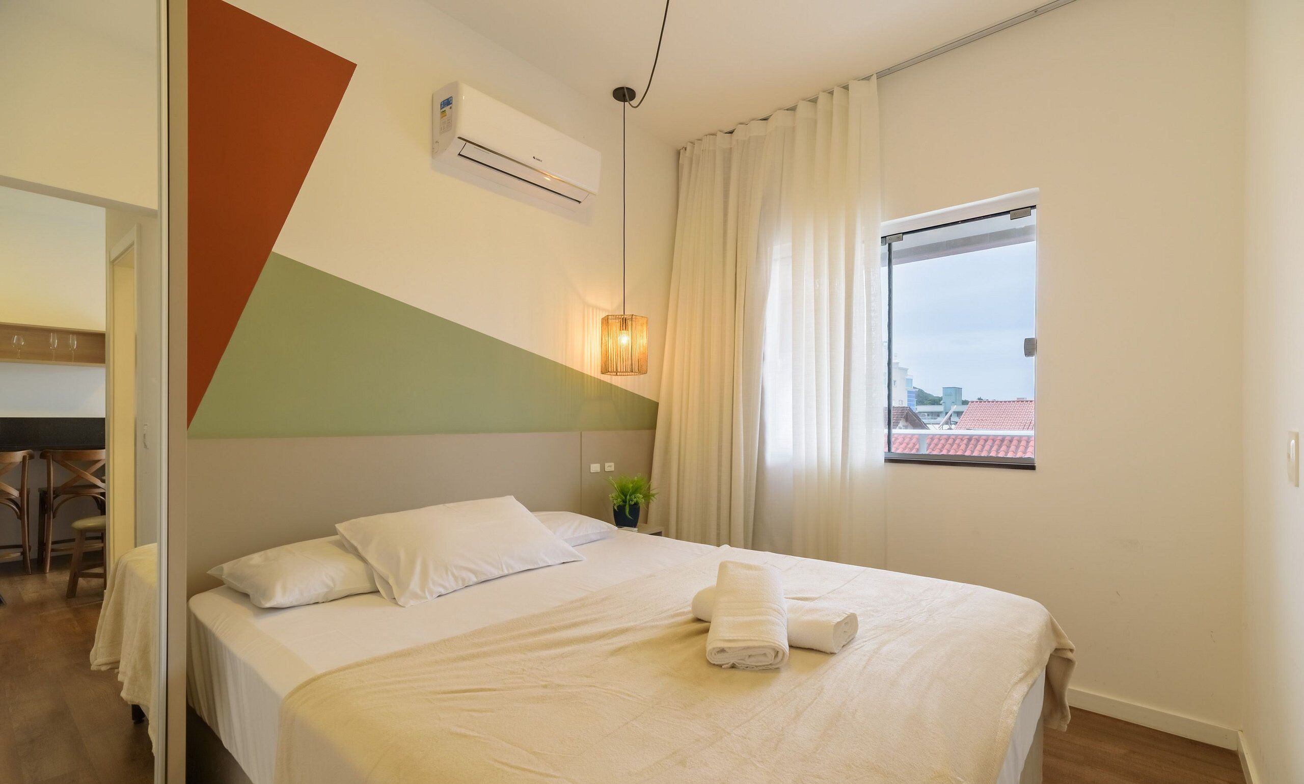 A hotel room with a bed, a window with curtains, an air conditioning unit, and a plant on the nightstand.