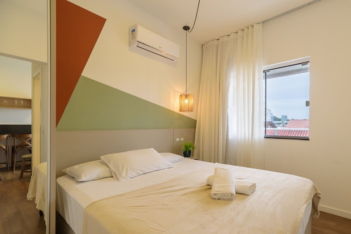 Ein Hotelzimmer mit Bett, einem Fenster mit Vorhängen, einer Klimaanlage und einer Pflanze auf dem Nachttisch.