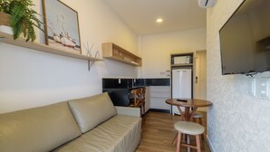 A compact living space with a sofa, a small dining table, a kitchen area with a refrigerator, and a mounted television. - Studio w/ Pool nr Beach Bombinhas Vec112 (Bombinhas)