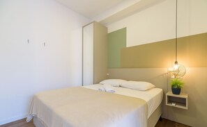 Un lit bien fait, avec du linge de lit blanc, une table de chevet agrémentée d’une plante et un luminaire fixé au mur.