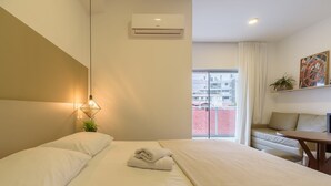 Ett hotellrum med en säng, ett skrivbord, en stol, en luftkonditionering, ett fönster med utsikt över byggnader och en växt.