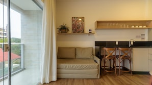 Ett kompakt vardagsrum med en soffa, två trästolar, ett litet köksutrymme med en svart bänkskiva och ett fönster med en gardin.