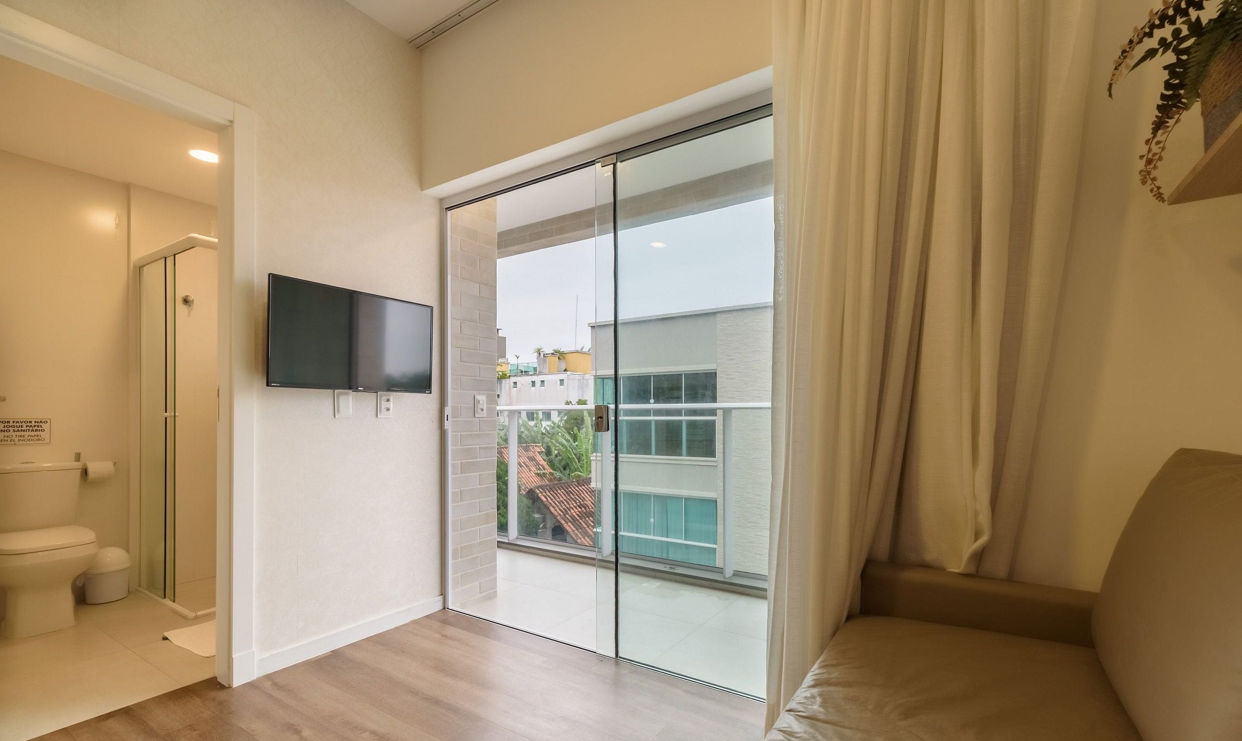 A hotel room with a sliding glass door to a balcony, a flat-screen TV mounted on the wall, and a toilet with a trash bin.