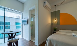 Quarto de hotel com cama, mesa de cabeceira com abajur, televisão na parede e varanda com mesa e cadeiras.