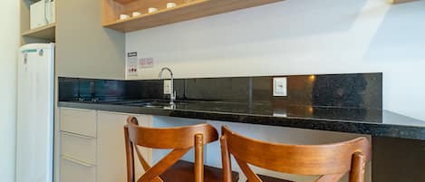 Una cocina con encimera negra, armarios blancos y un refrigerador blanco, además de dos sillas de madera.
