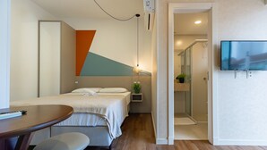 Habitación de hotel con cama, escritorio, silla, televisor y zona de ducha.