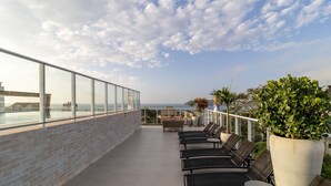 A rooftop terrace with a glass balustrade, lounge chairs, potted plants, and a view of the ocean. - Studio 350m From Beach Bombinhas Vec213 (Bombinhas)