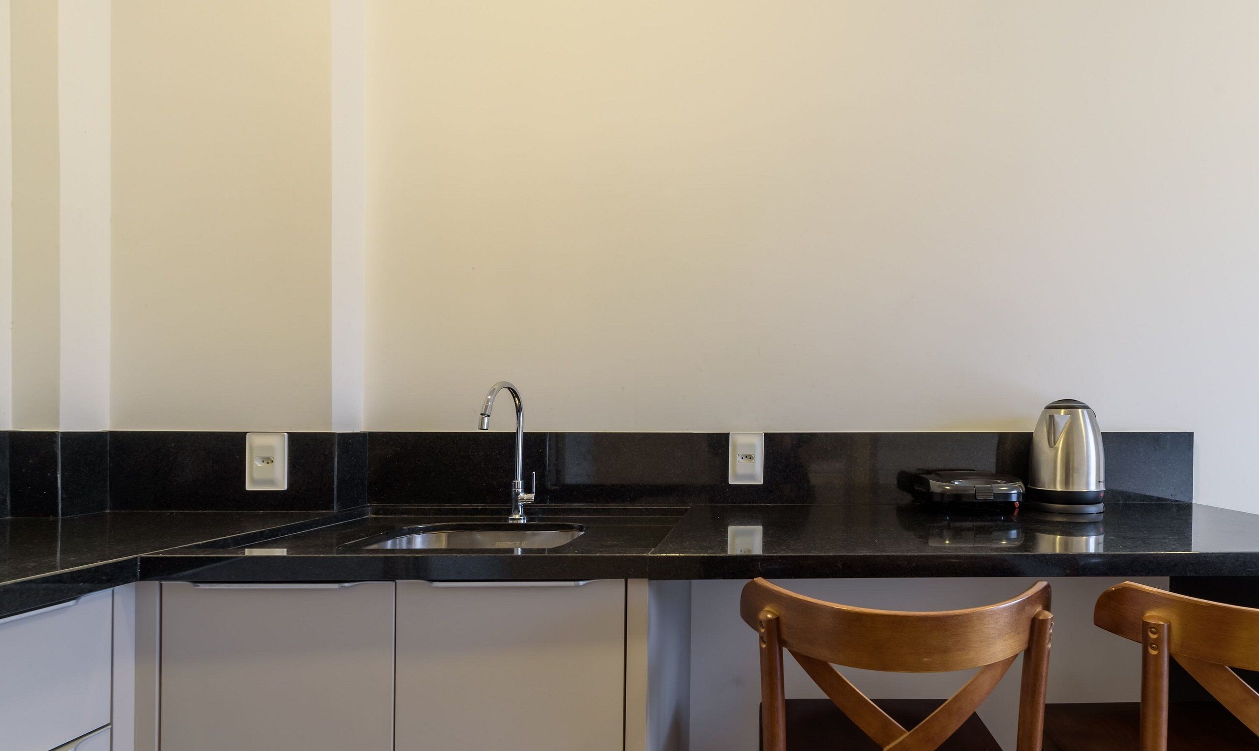 Una cocina con encimera negra, un fregadero de acero inoxidable y una tetera. Hay dos sillas de madera y un armario blanco debajo de la encimera.