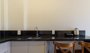 Eine Küche mit schwarzer Arbeitsplatte, einem Edelstahlspülbecken und einem Wasserkocher. Zwei Holzstühle und ein weißes Schranksystem befinden sich unter der Arbeitsplatte.