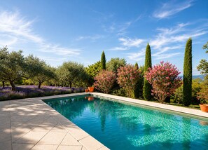 Ein Swimmingpool mit einer Fliesenveranda, umgeben von verschiedenen Bäumen und blühenden Pflanzen.