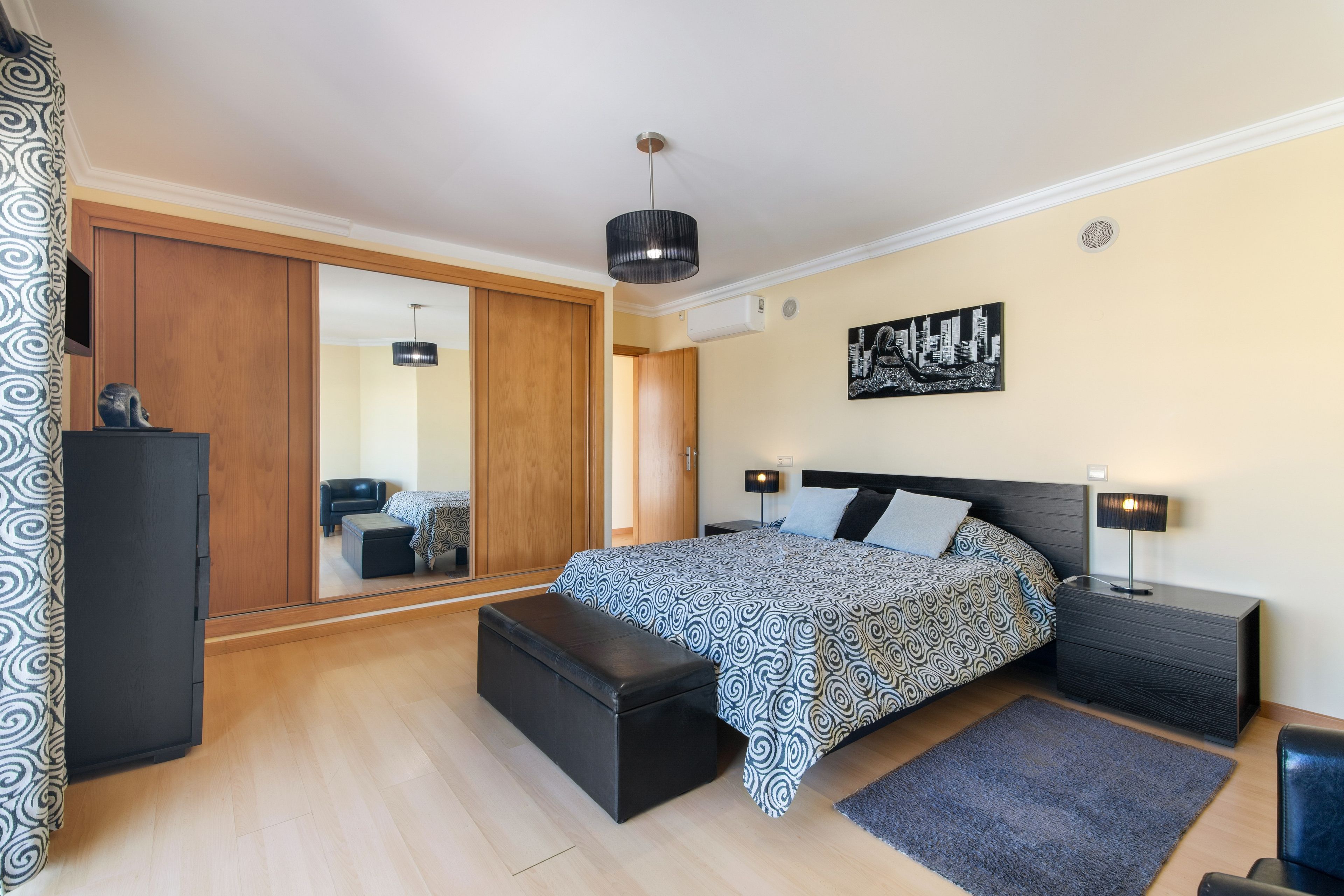 Un dormitorio con cama, mesitas de noche, un armario, un espejo y una obra de arte con un paisaje urbano en la pared.