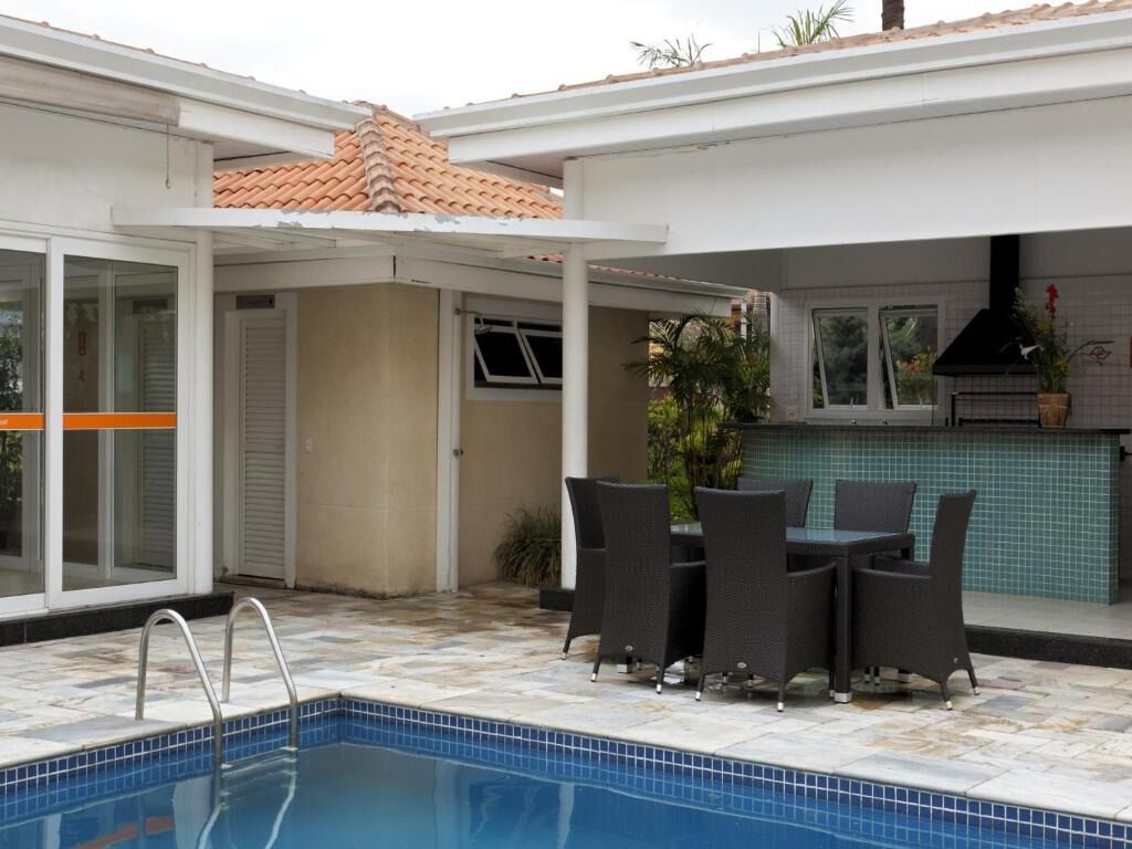 Piscina com deck revestido, varanda coberta com mesa e cadeiras, e área de cozinha com painel atrás do fogão.