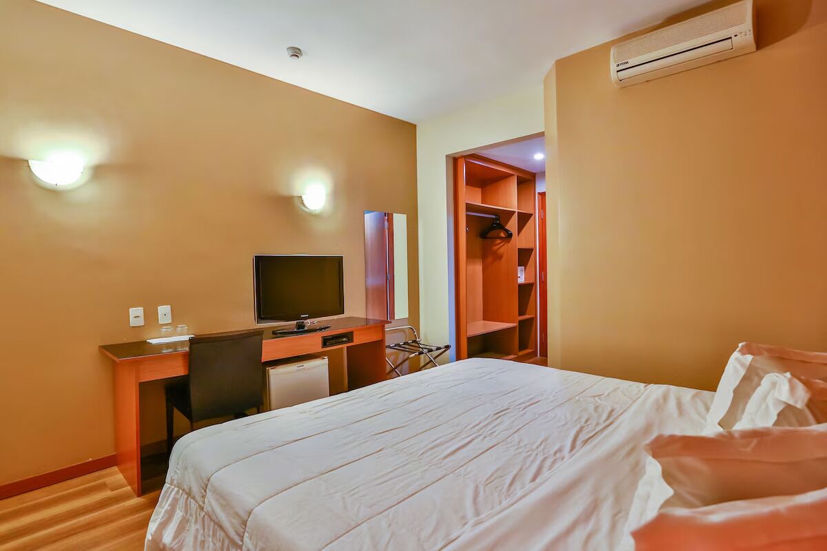Quarto de hotel com cama, escrivaninha com computador, televisão e guarda-roupa.