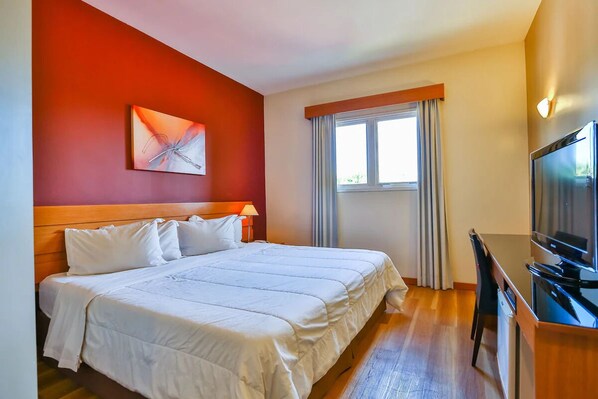 A hotel room with a bed, a desk with a TV, a window with curtains, and a painting on the wall. - Bright Room | Near São Paulo Business Hub | Close to Jardim Botânico (São Paulo)