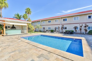 Ein Pool mit Fliesen umgeben, eine überdachte Terrasse und ein Gebäude mit rotem Dach und mehreren Fenstern.
